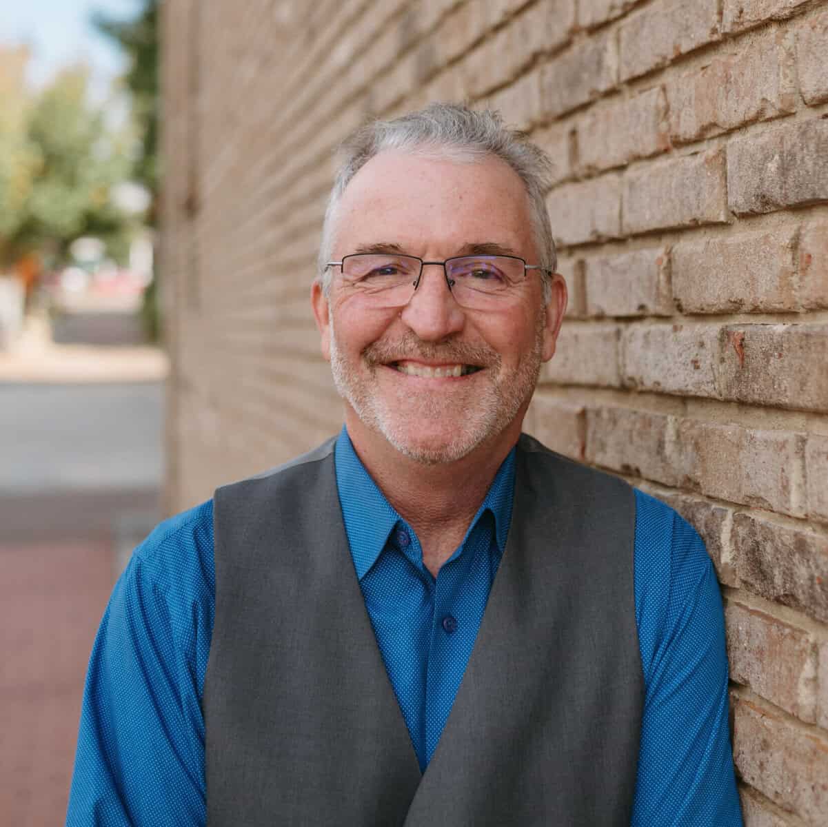 Smiling mature man in blue shirt and vest standing by brick wall outdoors.