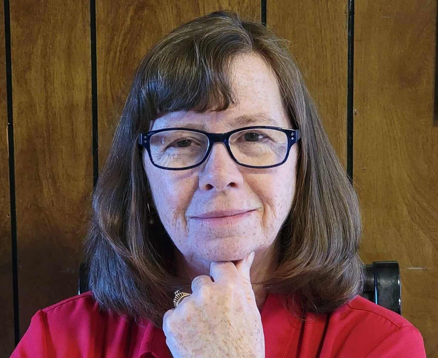 Woman with glasses smiling, wearing a red top, in a warm wooden setting.