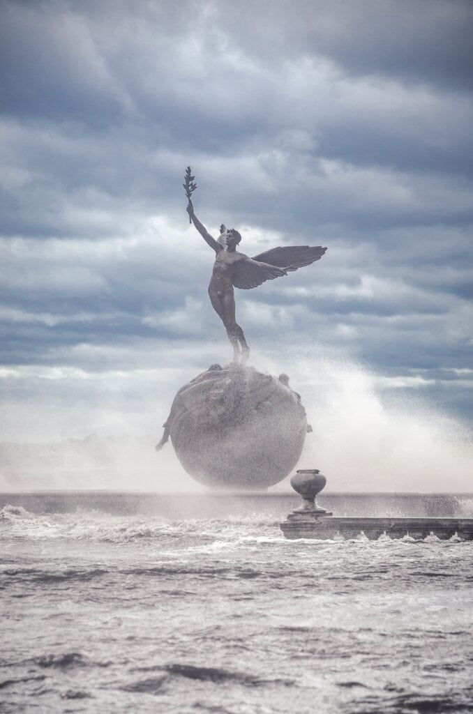 Artistic fountain sculpture of a woman holding a torch, set against cloudy sky.