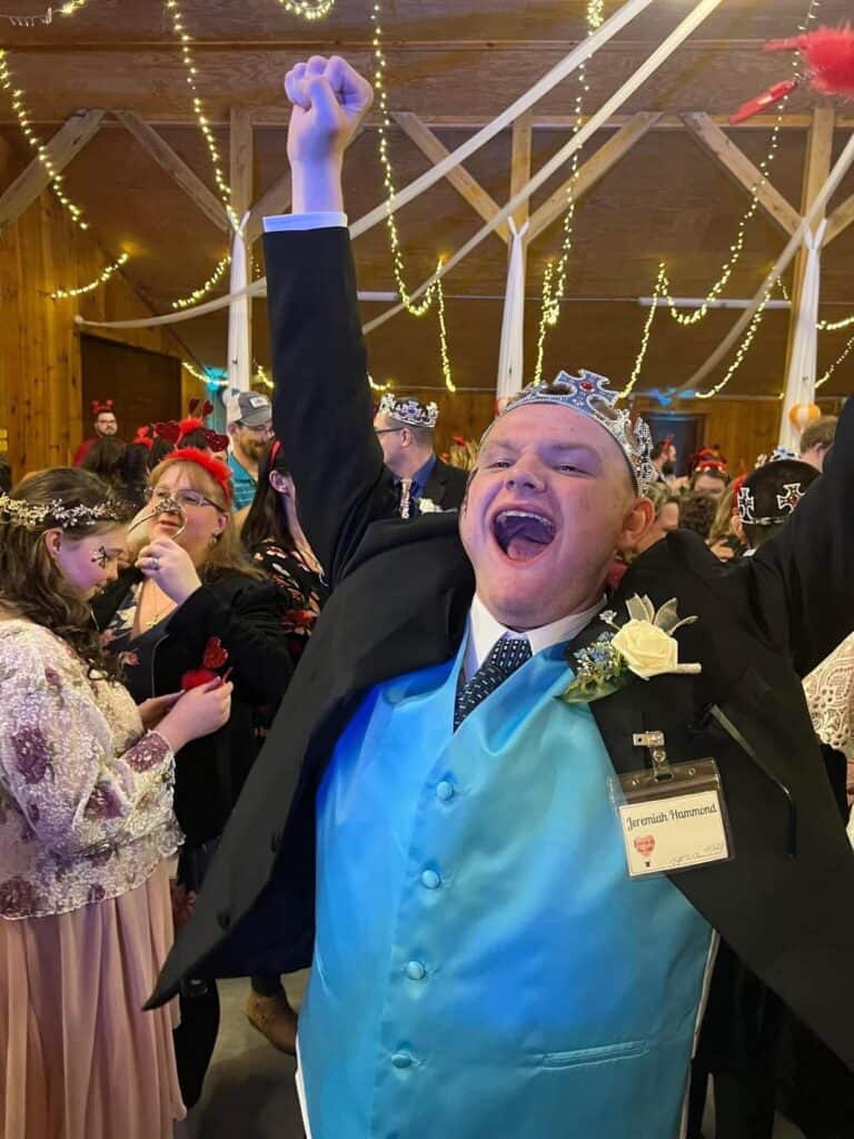Joyful young man celebrating at Night to Shine event with crown and tuxedo.