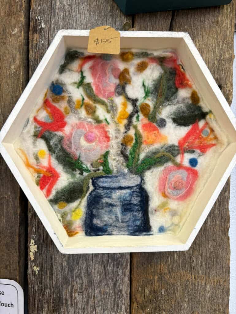 Wool felted art piece of a vase with flowers in a hexagon frame.