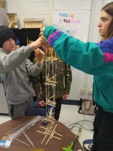 Students building a tall structure with wooden sticks in classroom setting.