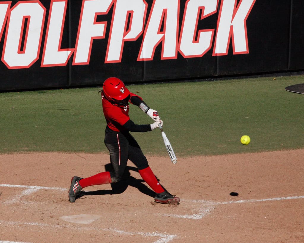 FHS athlete in red uniform swinging bat at baseball, preparing to hit the ball.