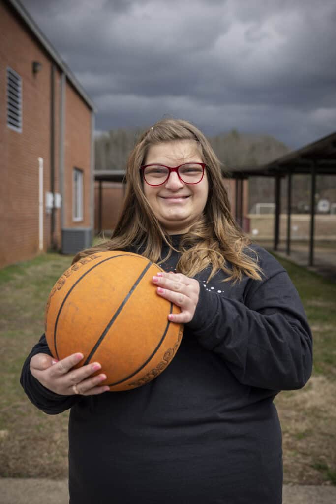 Young girl holding basketball outdoors on cloudy day at school.