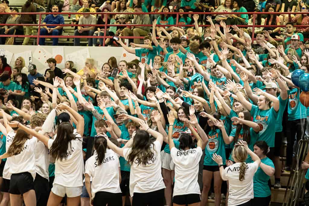 Packed gymnasium during FHS March Madness event with students cheering and raising hands.