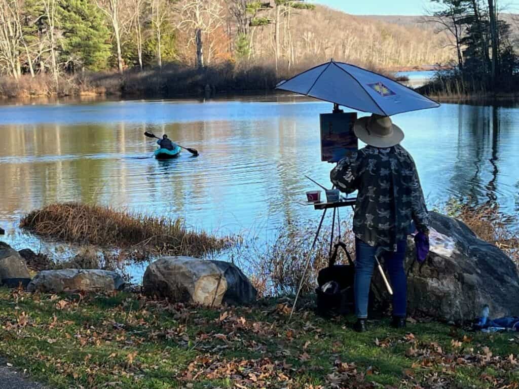Artist Carol Conti creating art outdoors by a serene lake scene.