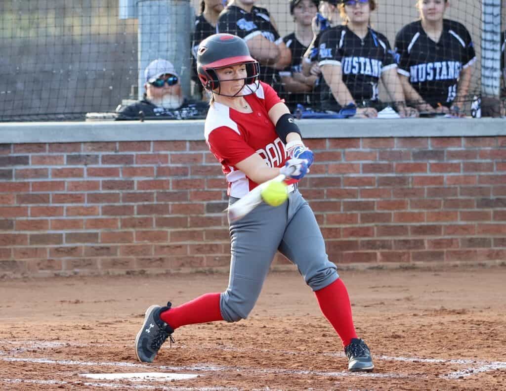 Female softball player swinging at a pitch during a game, wearing red and gray uniform with helmet.