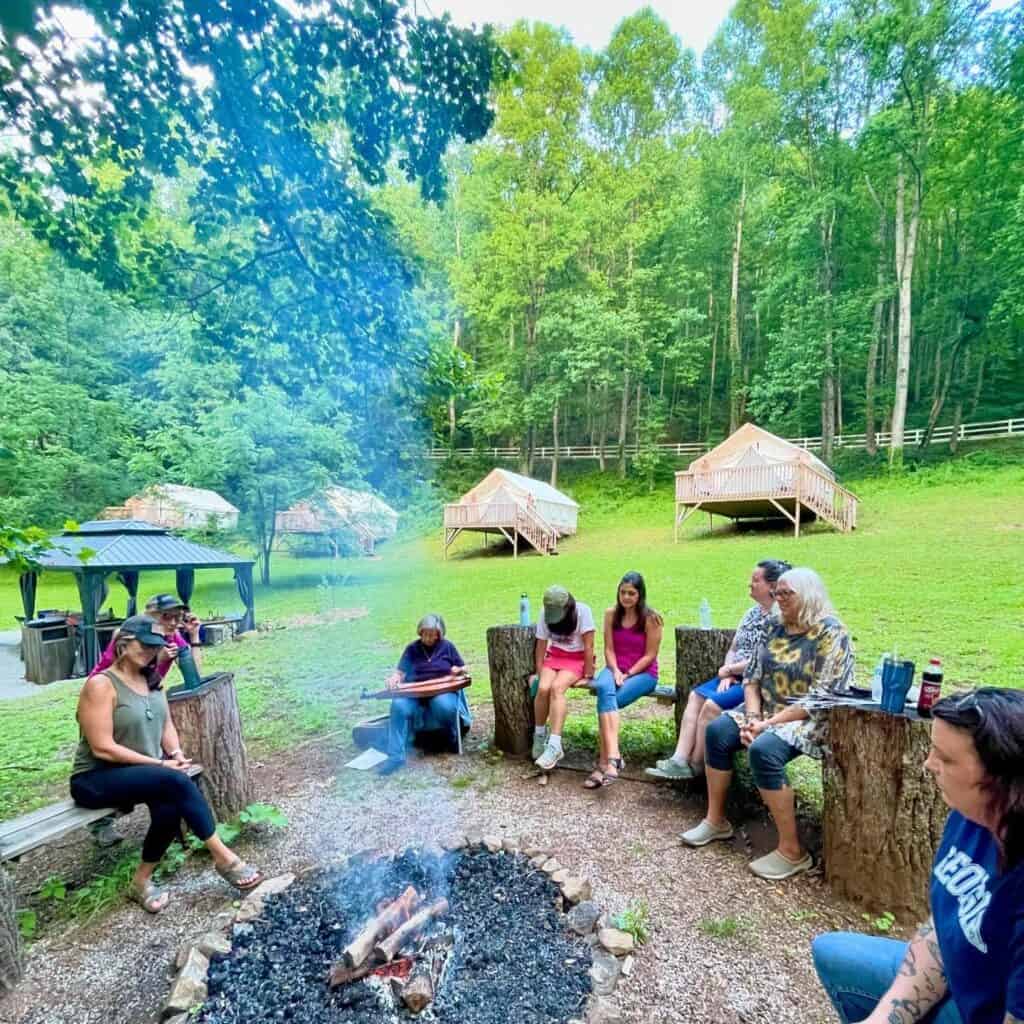 Outdoor summer camp gathering with people sitting around a campfire in a forest setting.