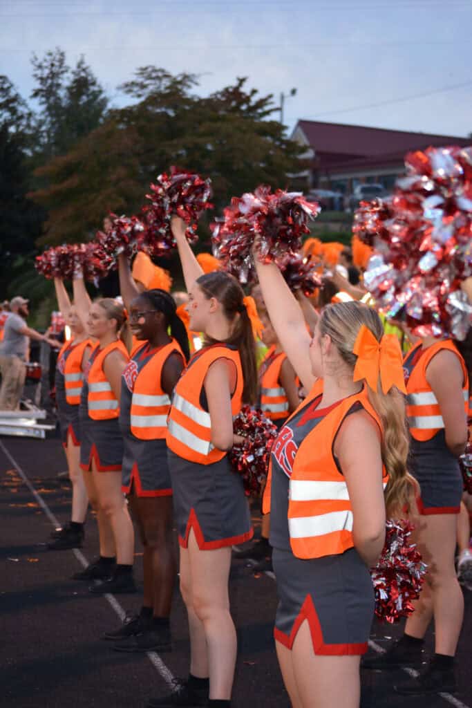 Cheerleaders in orange safety vests performing at a cross country race event.