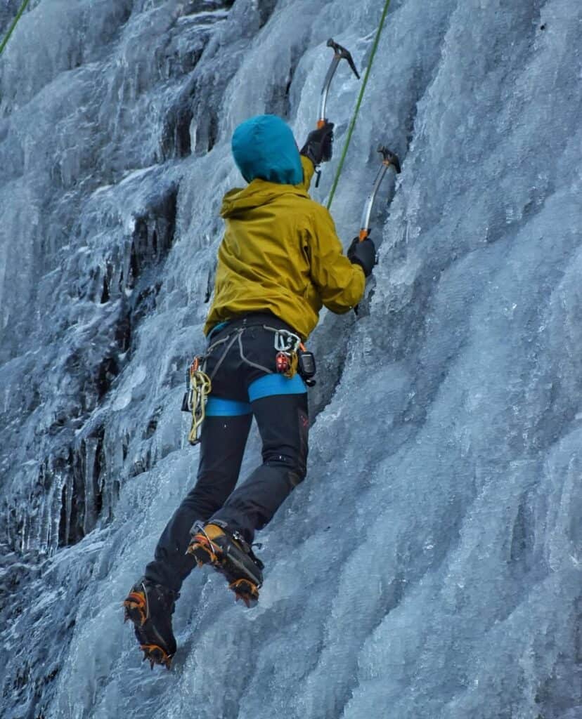 Ice climber on frozen waterfall with ice axes and crampons for winter climbing.