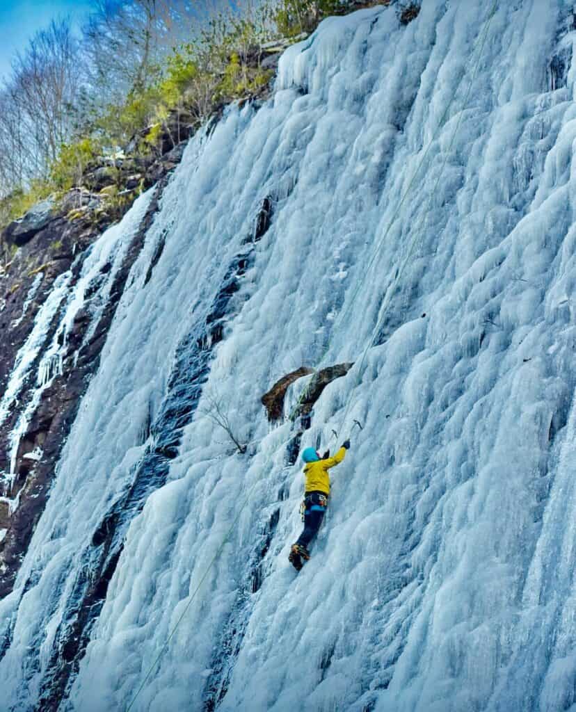 Ice climber scaling icy waterfall with safety gear in winter.