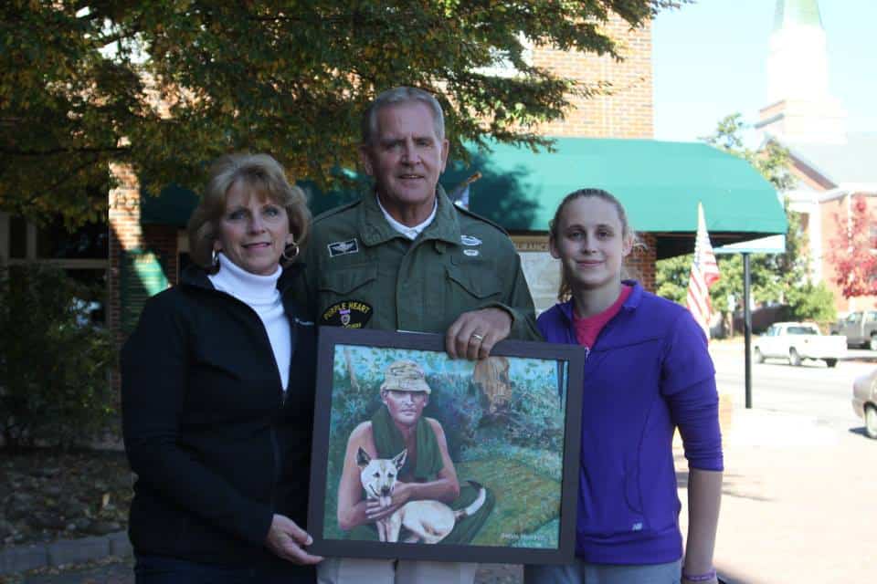Soldier holding a framed photo with two women outdoors, Vietnam jungle background.