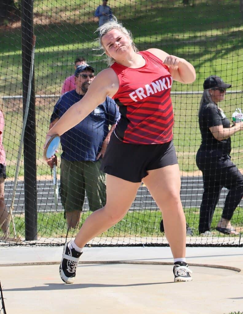 Female athlete in red and black uniform throwing discus at track meet.