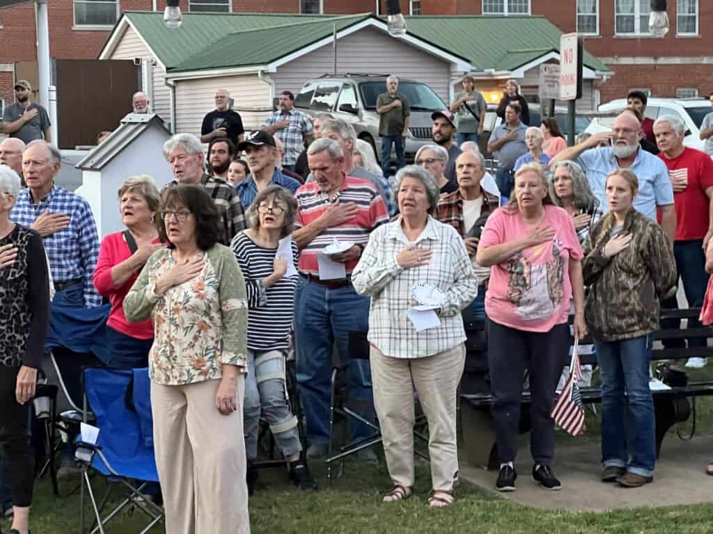 People gathered outdoors, holding American flags, praying during a community event.