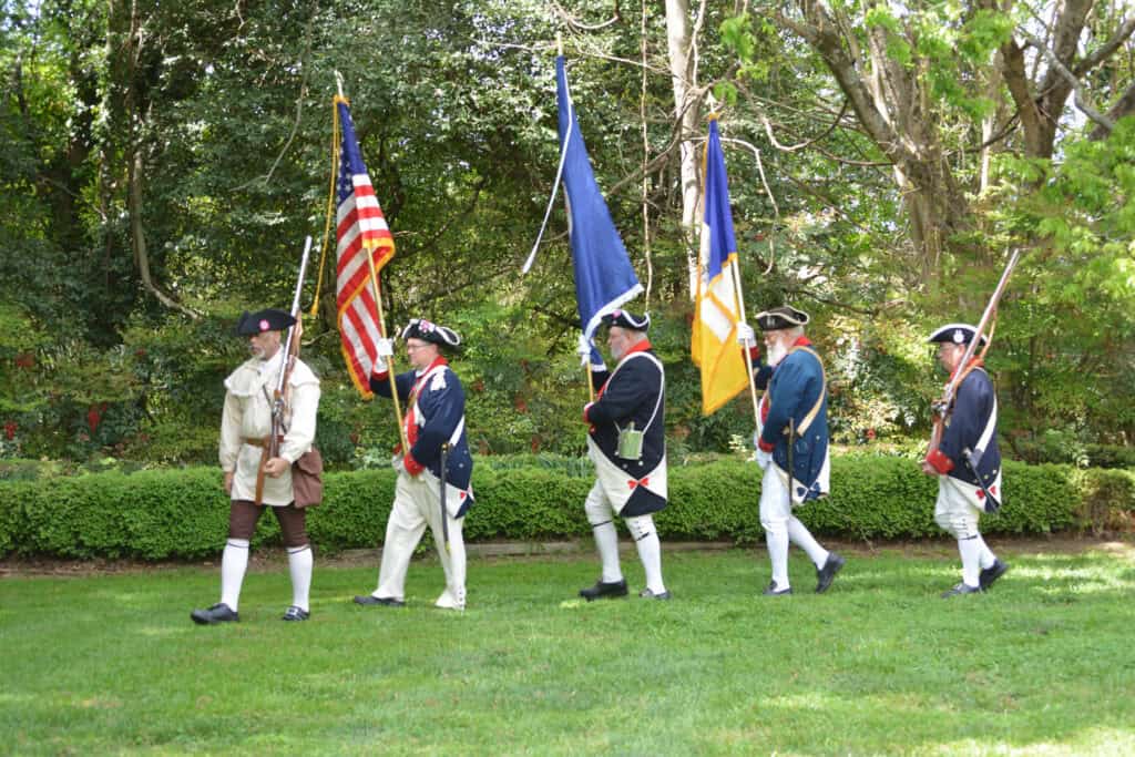 Historical reenactors in Revolutionary War uniforms carrying American flags.