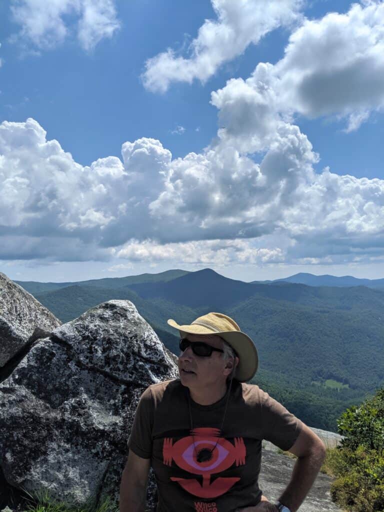 Hiker wearing a hat and sunglasses enjoying a mountain landscape with clouds and blue sky.