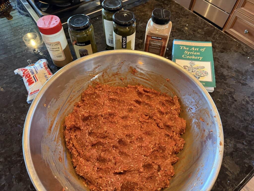 Raw meat mixture in a stainless steel bowl with spices and cookbook in background.