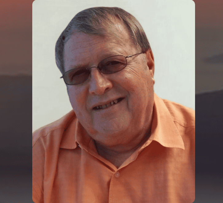 Portrait of John Paul Barbee smiling, wearing glasses and an orange shirt.