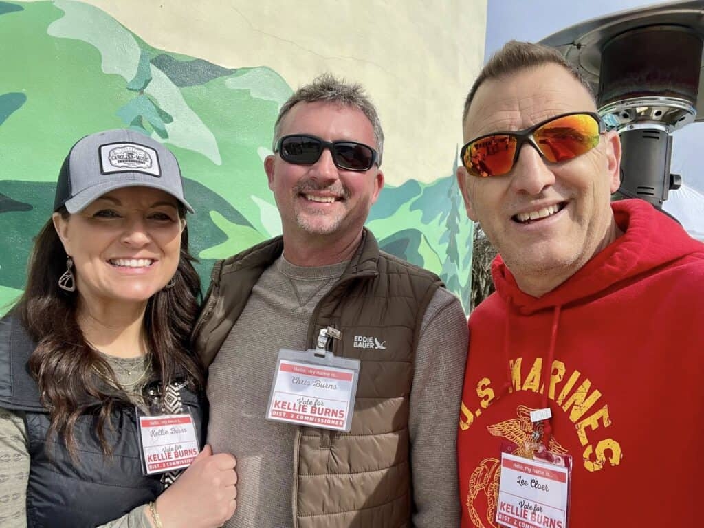 Group of three smiling people at outdoor event, wearing name tags and casual clothing.