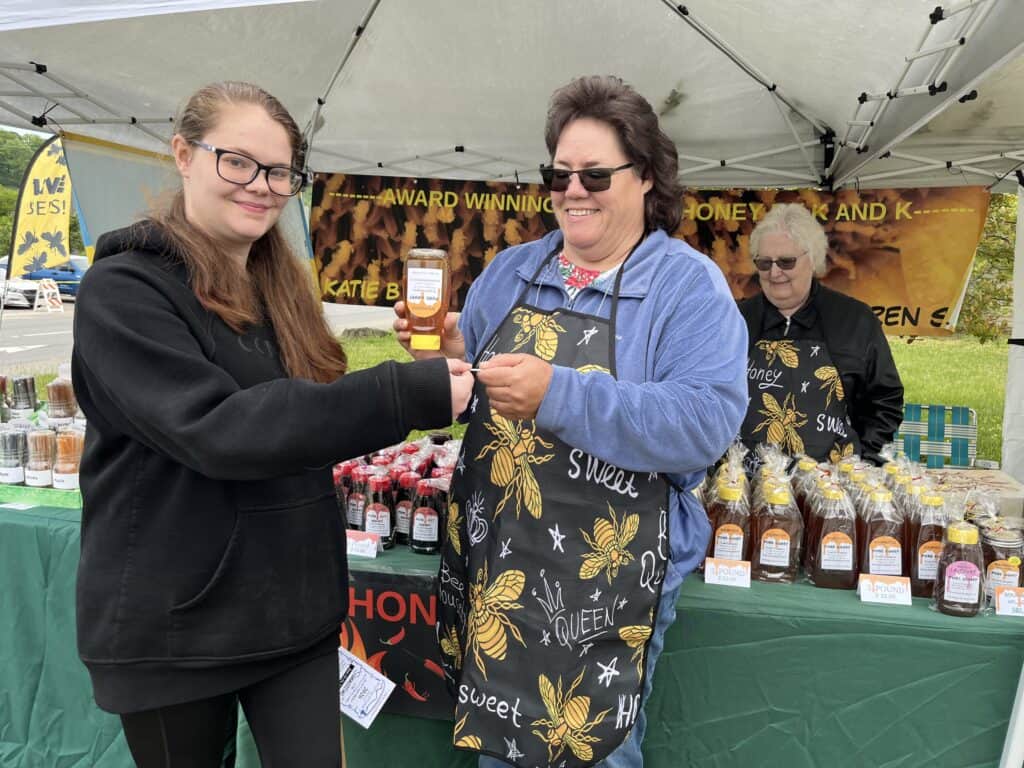 Women exchanging honey jars at spring fair vendor booth in outdoor market.