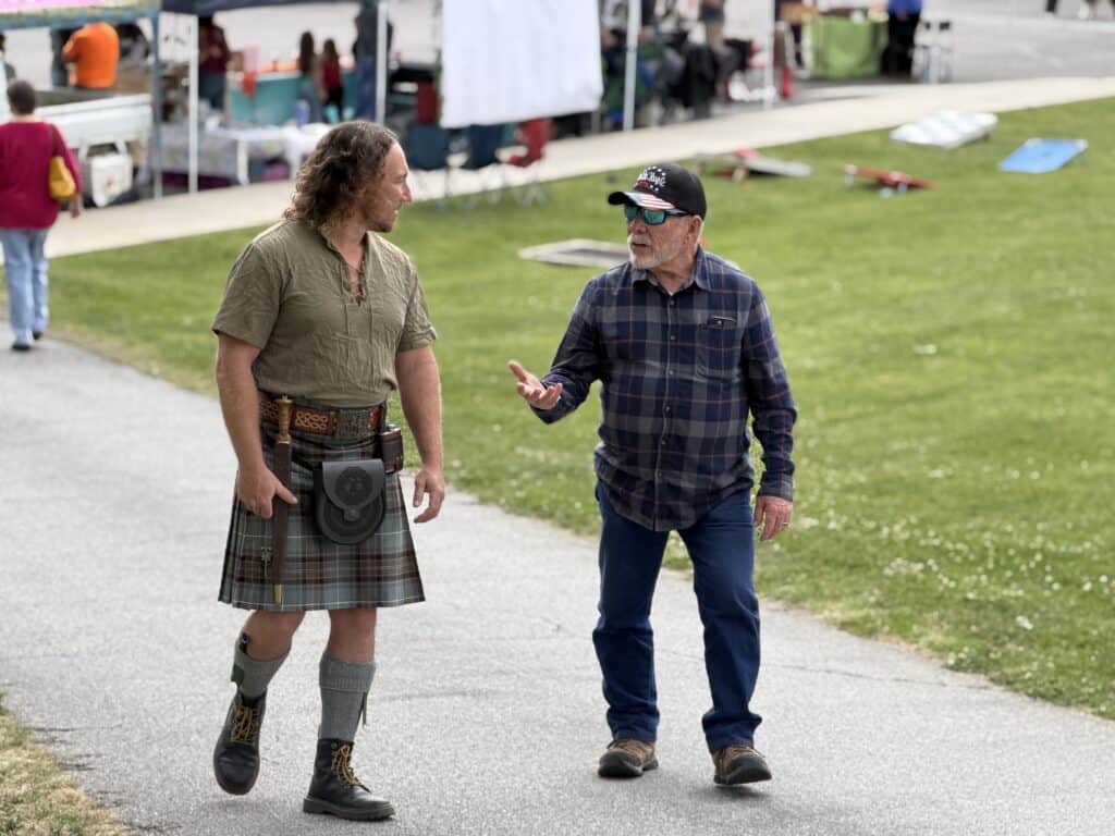 Two men conversing at outdoor market with tents and boats in background.