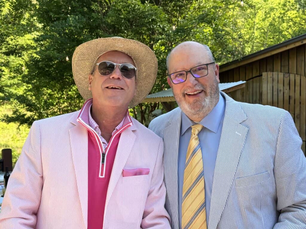 Stylish men in suits smiling outdoors during daytime, one wearing a hat and sunglasses, with lush gr.