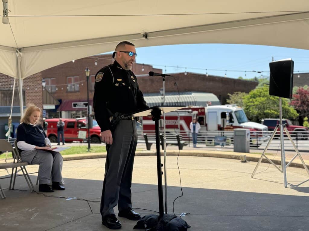 Police officer speaking at outdoor community event, wearing uniform and sunglasses.