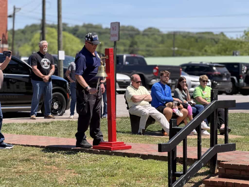 Man ringing a bell at a memorial service with seated attendees outdoors.