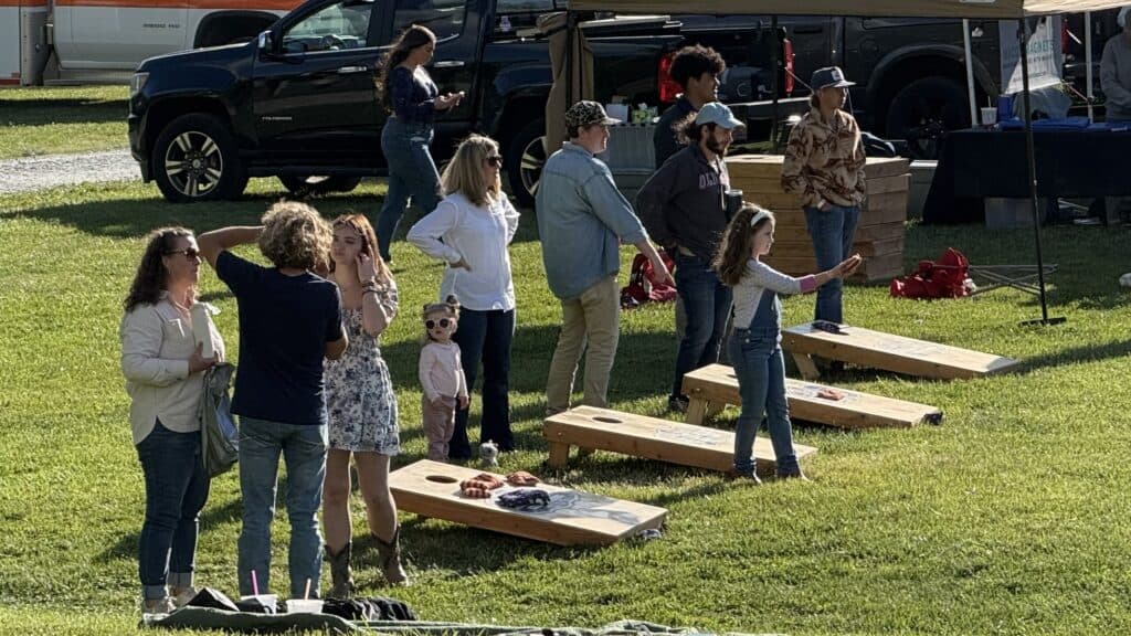 Children playing cornhole at a spring festival with families and vendors in the background.
