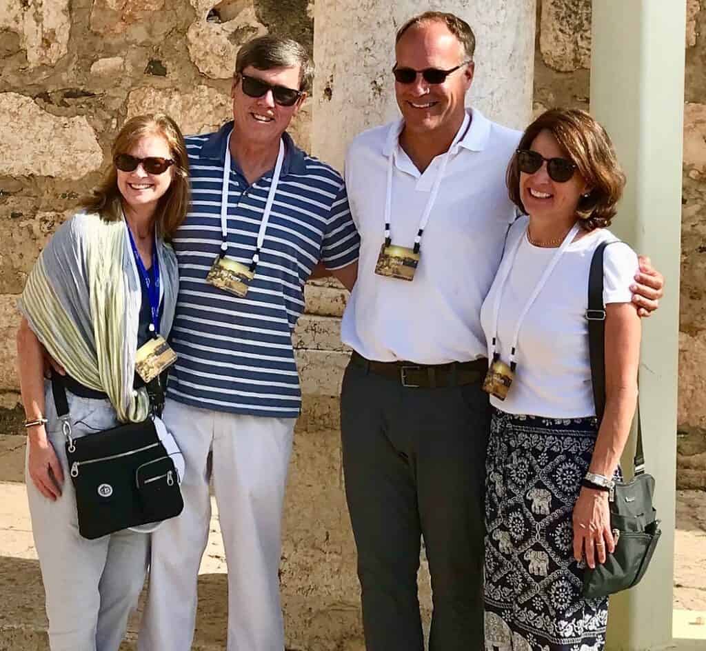 Group of four smiling adults at outdoor event, wearing sunglasses and casual attire.