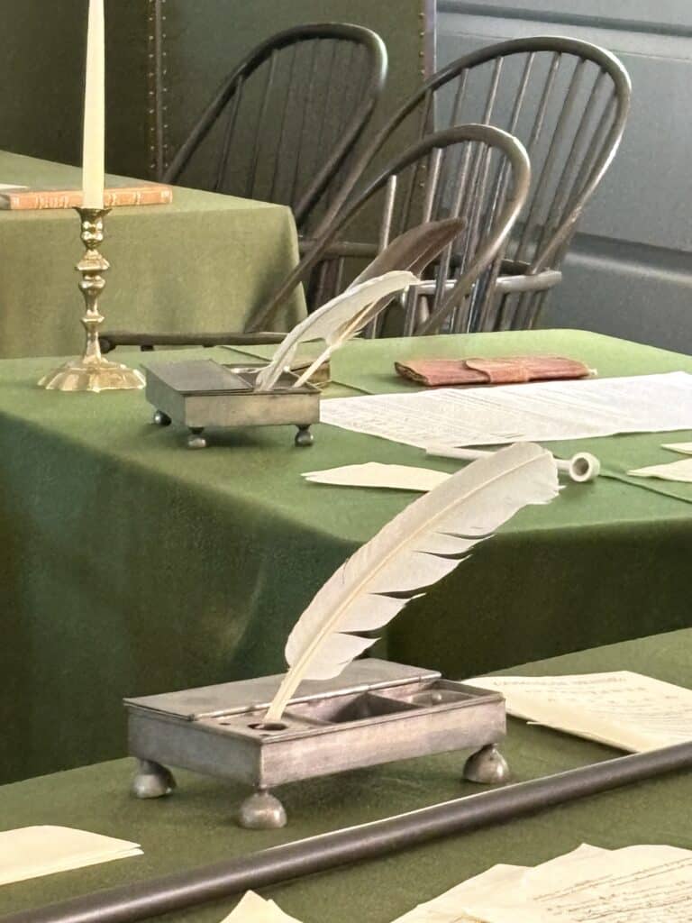 Vintage quill and ink set on a green table with chairs in background.