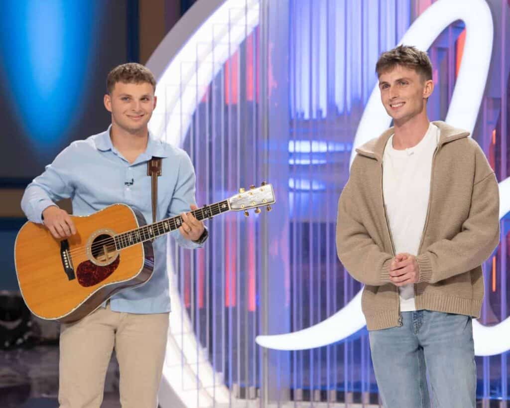 Two young men on stage, one playing guitar, smiling and engaging with the audience.