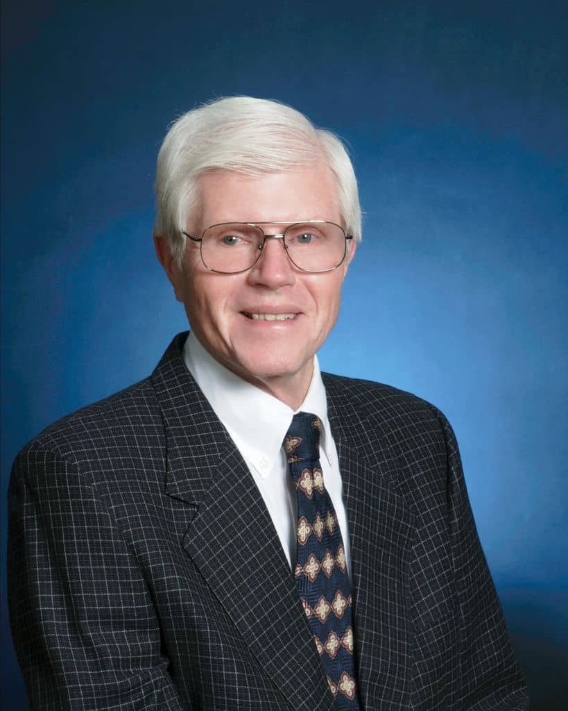 Senior businessman in formal suit with glasses, smiling against blue background.