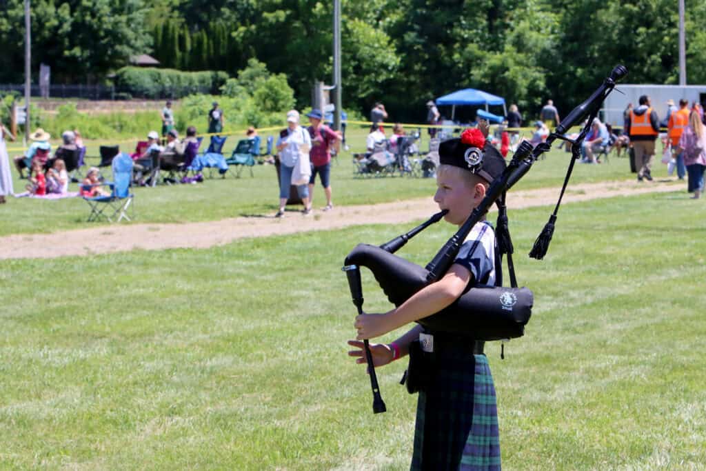Child playing bagpipes at outdoor festival with crowd and greenery.
