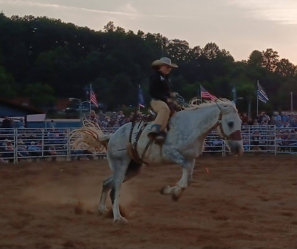 Rodeo cowboy riding a white horse at sunset with American flags in background.