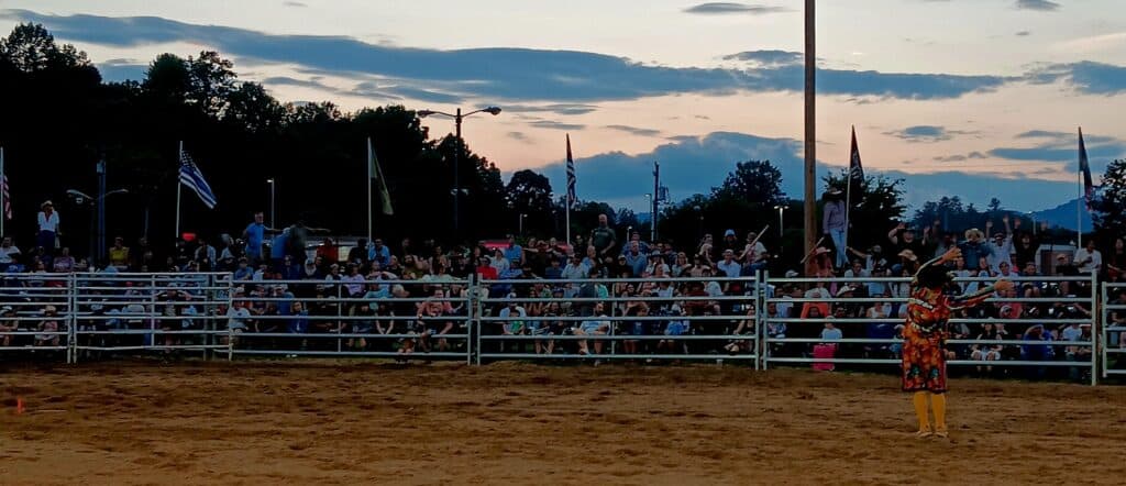 Crowd watching a rodeo event at sunset with flags and fencing.