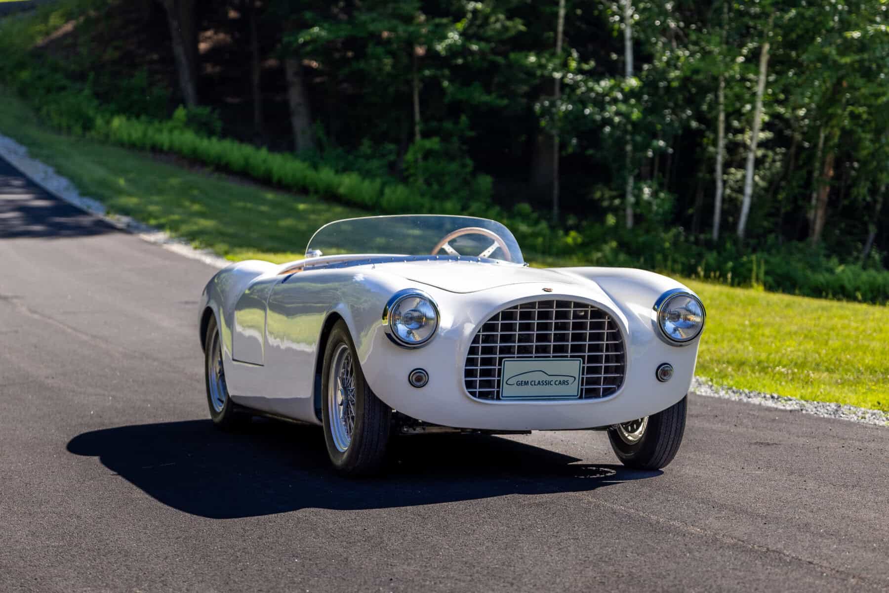 Vintage white sports car driving on a scenic road with trees in the background.