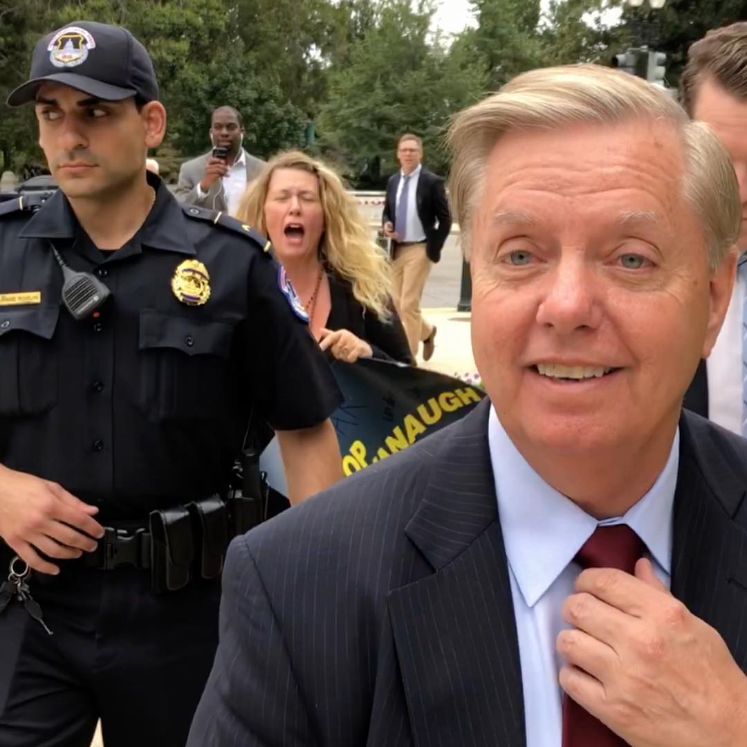 Lindsey Graham smiling at a public event with police and protesters in background.