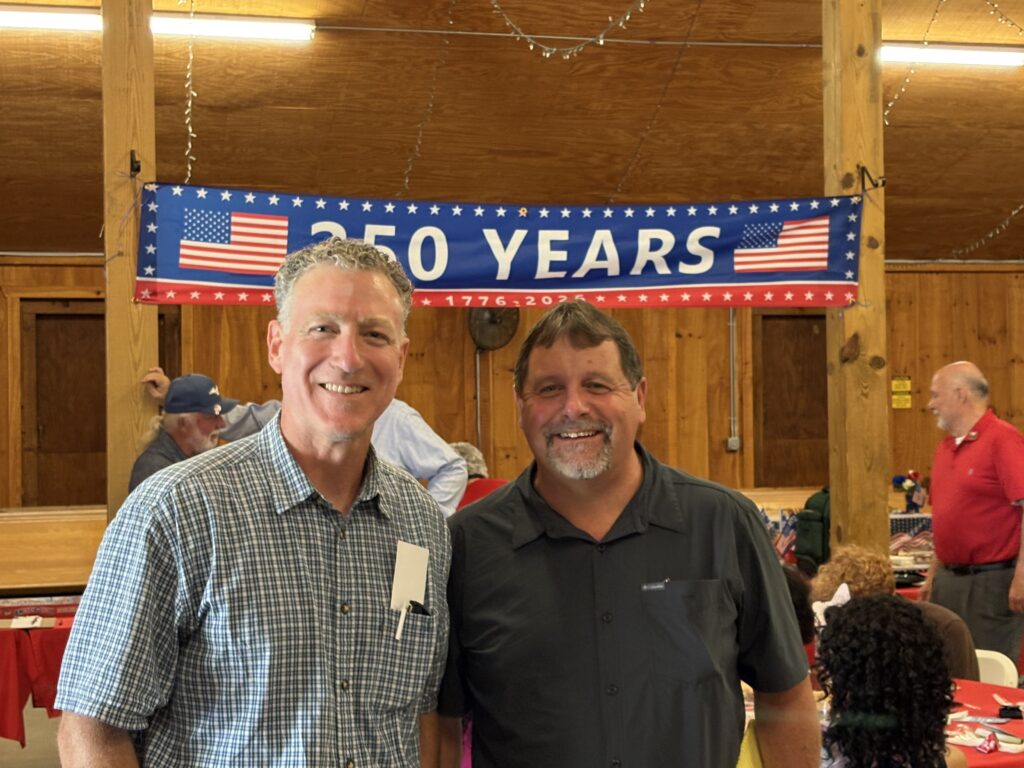 Two men smiling at a GOP event with a 250 Years banner in the background.