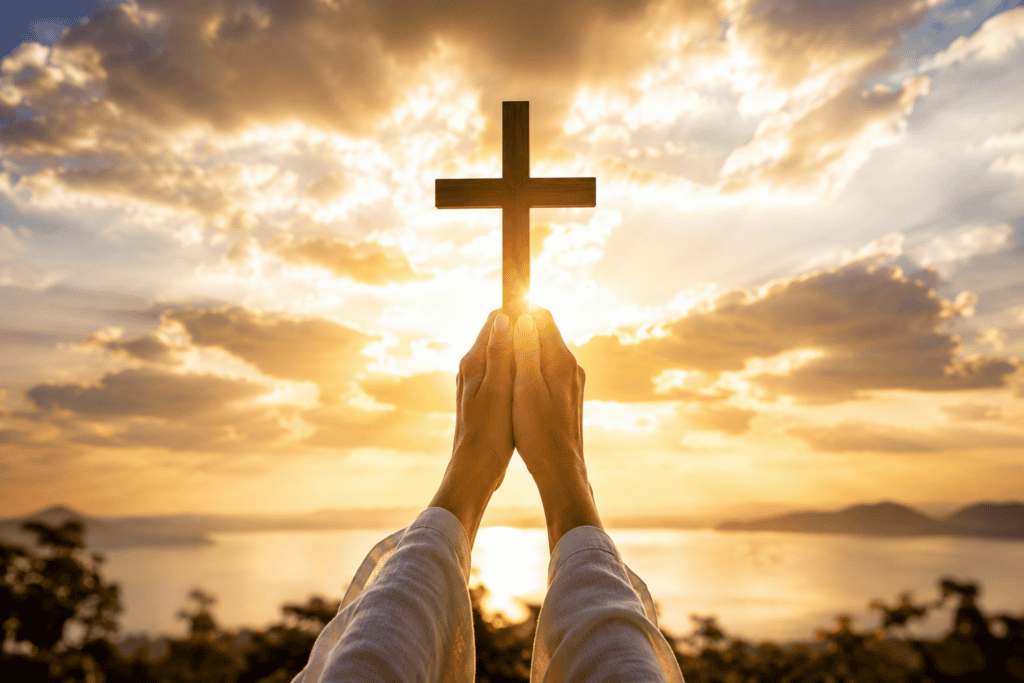 Person holding a cross against a sunset sky with clouds and water.