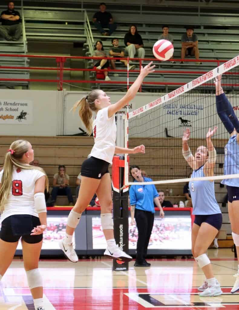 Girls playing volleyball during a school match with spectators in the background.