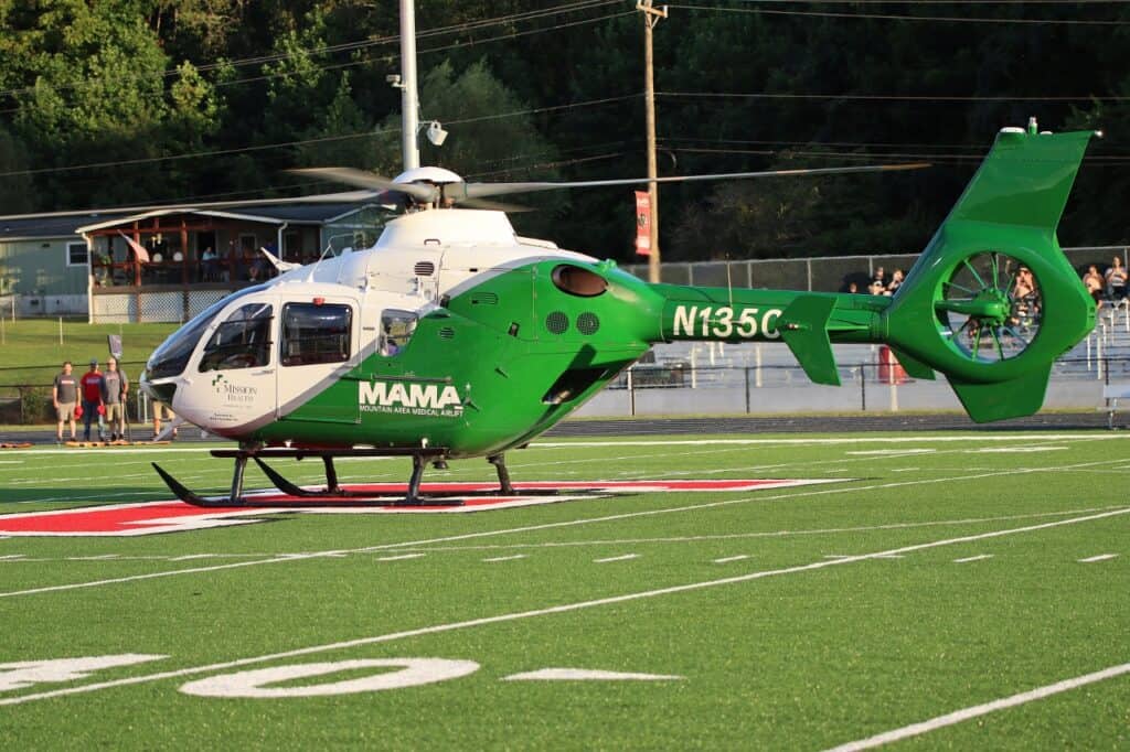 Green and white helicopter on football field with spectators in background.