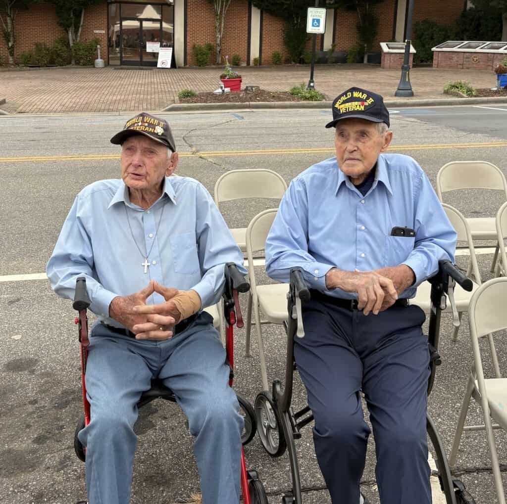 Two elderly veterans in uniform hats sitting outdoors for a celebration.