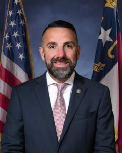 Professional portrait of a man in a suit with American flags in background.