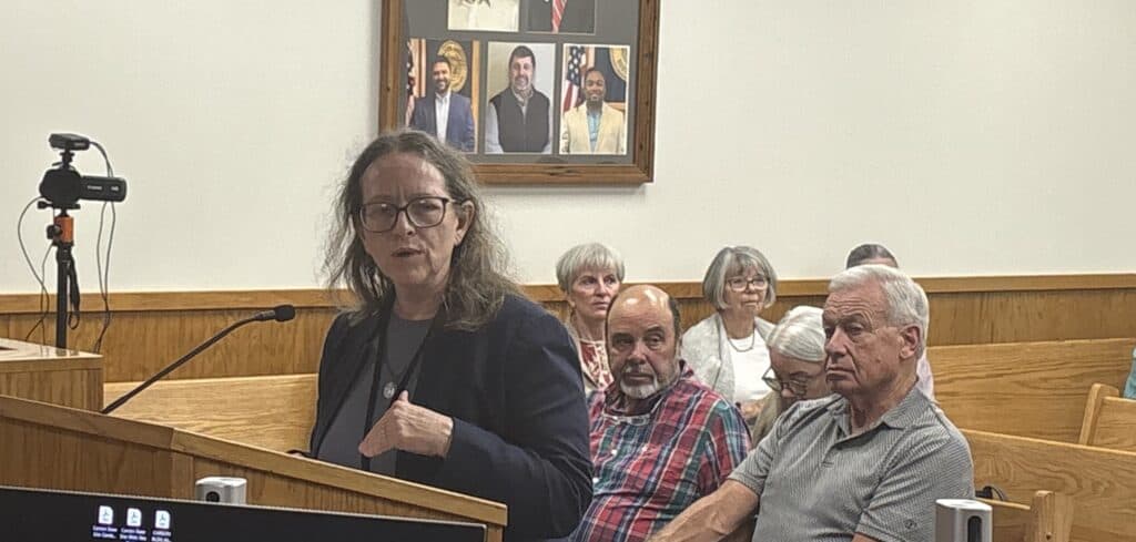 Woman speaking at a public meeting with seated audience members.