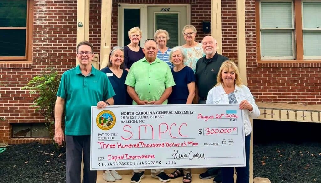 Group of people holding a large check outside a brick building.