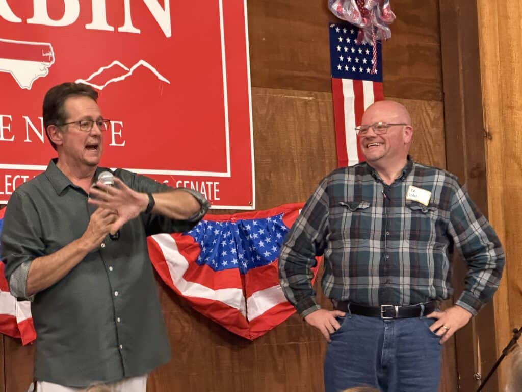Two men speaking at a political event with American flags and patriotic decorations.
