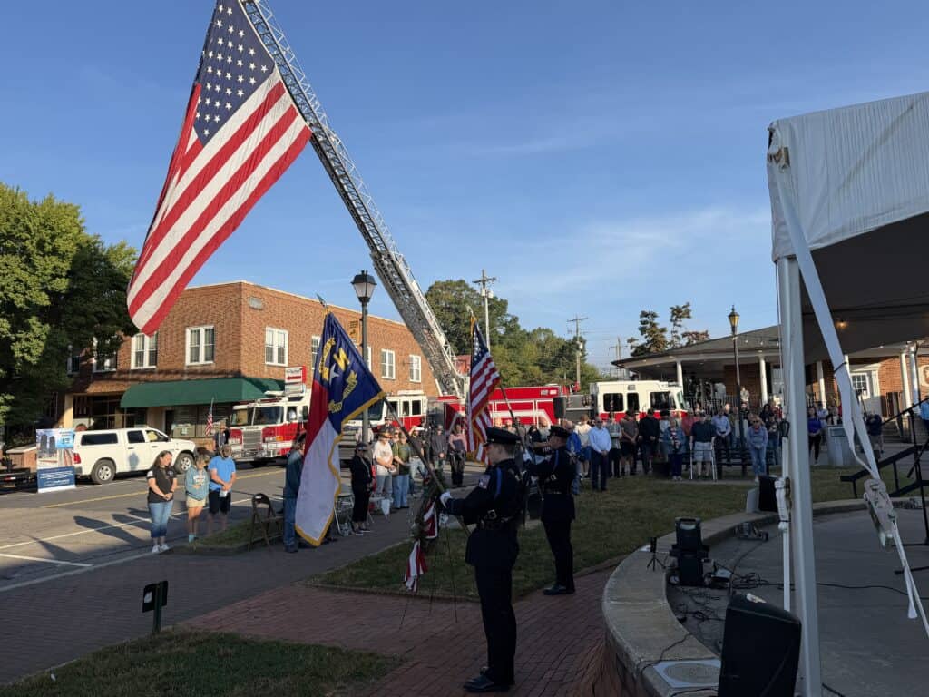 Firefighters and community gather for memorial event with American flag and fire truck.