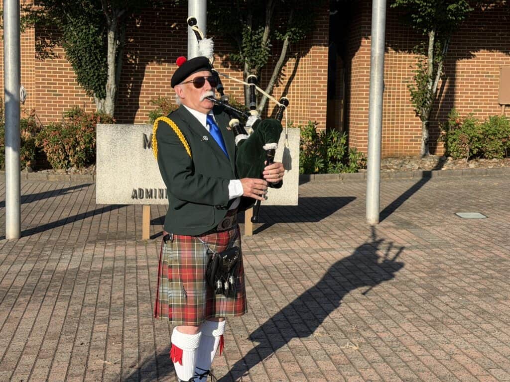 Scottish bagpiper wearing kilt and playing bagpipes outdoors.