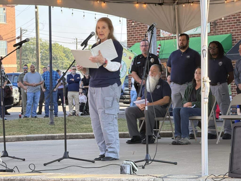 Woman speaking at outdoor community event with audience and officials.